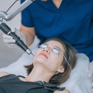 žena leži na medicinskom krevetu u dermatološkoj ordinaciji, a iznad nje stoji dermatolog u plavoj uniformi koji drži laser pored njenog lica - u toku je lasersko lečenje rozacee