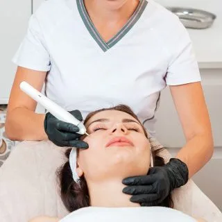 žena zatvorenih očiju leži na kozmetičkom stolu u dermatološkoj ordinaciji dok lekarka drži laser na njenom licu - u toku je lasersko uklanjanje fleka i hiperpigmentacija