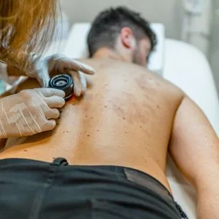 muškarac leži na stomaku u dermatološkoj ordinaciji, a dermatolog pregleda kožu na njegovim leđima
