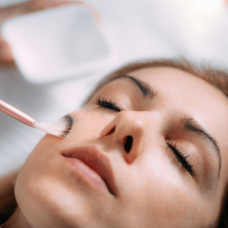 žena zatvorenih očiju leži u dermatološkoj ordinaciji dok dermatolog na njenom licu izvodi hemijski piling