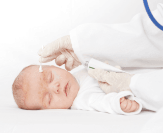 beba na pregledu kod dečijeg dermatologa: novorođenče u belom leži zatvorenih očiju, na obrvi je trag kreme, a iznad nje dečiji dermatolog koji vrši pregled