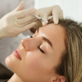 žena leži a ruke dermatologa u belim rukavicama drže špric i u glavu ubrizgavaju hair filer